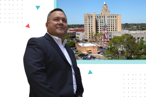 Business coach in Monroe, Louisiana, standing confidently in a suit with a cityscape of Monroe's downtown buildings in the background
